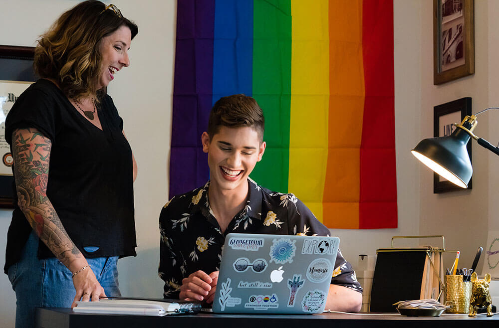 Young man on laptop talking to woman standing next to him with rainbow flag behind them