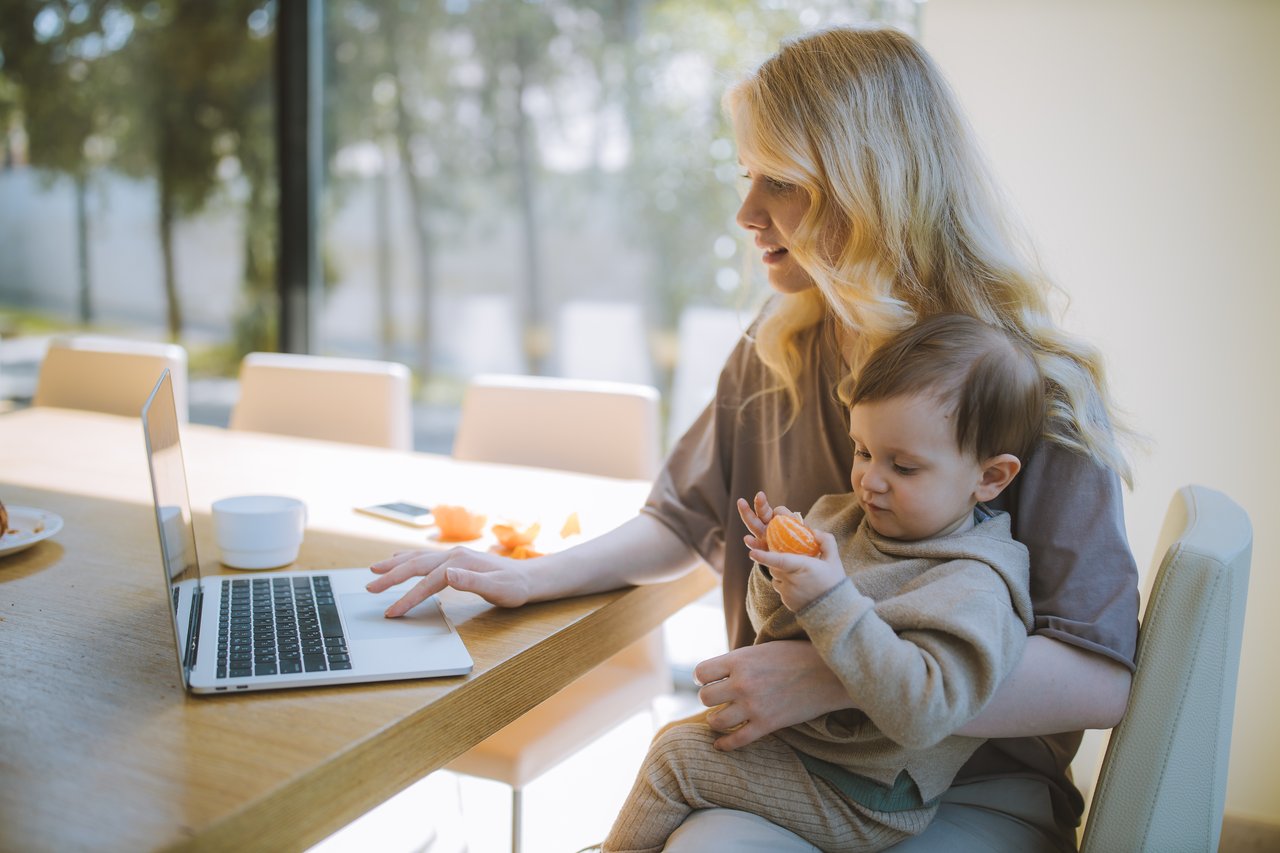 A mother sits at her computer with a toddler on her lap.