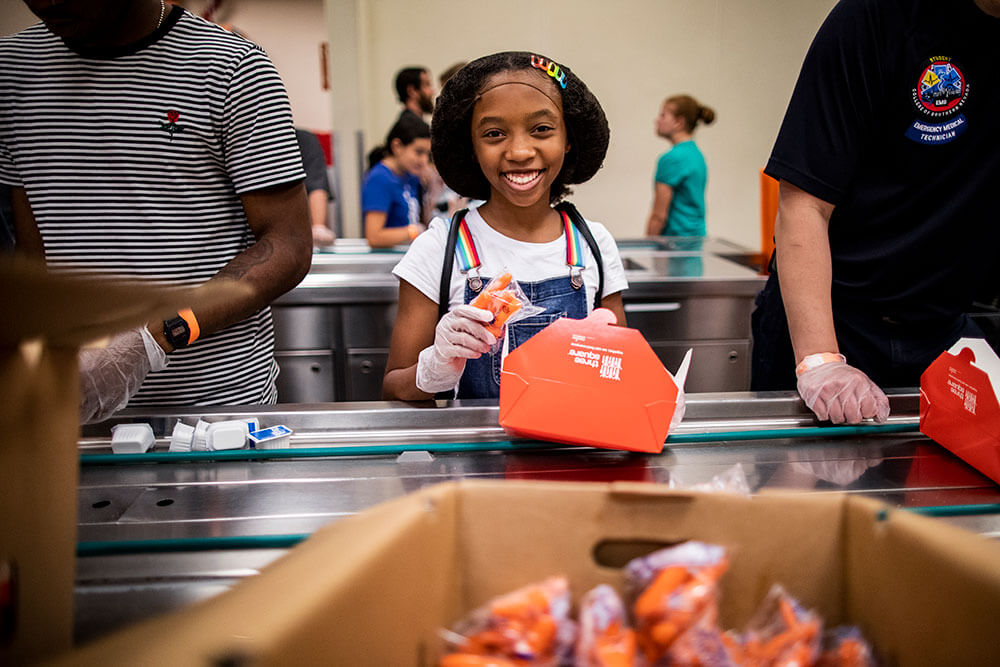 A young girl volunteer helps prepare food boxes in an assembly line.