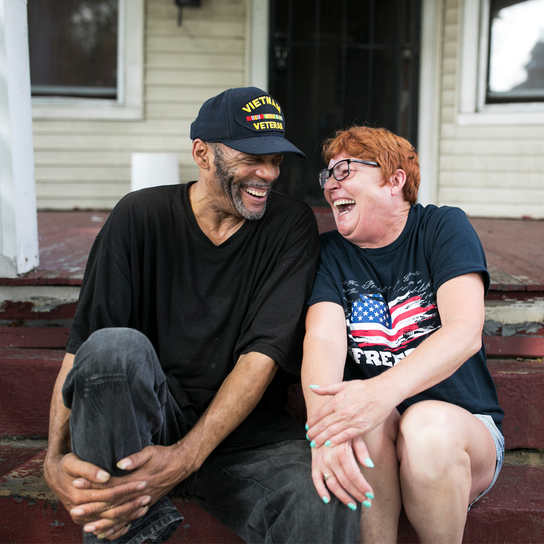 a man with a veteran hat on and a woman with an American flag shirt sitting on front porch stairs laughing